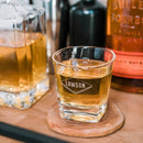 A short, heavy-bottomed Whiskey Glasses: Heirloom etched with the name LAWSON inside a diamond shape, filled with amber liquid and resting on a wooden coaster.