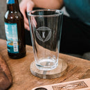 A clear glass from The Heirloom pint glass set, featuring a custom triangle-engraved letter K, sits on a wooden coaster next to a beer bottle.