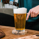 Beer being poured from a bottle into a Pint Glasses - Set of 4: The Heirloom personalized with a letter K logo on a wooden coaster.