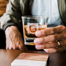 A person holds a Whiskey Essentials Bundle whiskey glass engraved with the letter E above a two-toned wooden and stone coaster.