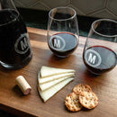 A Swanky Badger Wine Decanter - The Medalist and two stemless wine glasses, each etched with the letter M, sit on a wooden board with a wine cork, cheese slices, and crackers as part of a custom wine gift set.