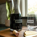 A personalized Wine Decanter - The Classic labeled Jackson sits next to a matching engraved wine glass labeled Rachel on a wooden board with a wine cork and cheese slices.