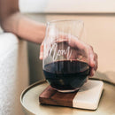 A hand holds a Wine Decanter - The Heirloom, a stemless wine glass engraved with Mom filled with red wine, resting on a two-toned wooden and marble coaster.