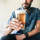 A man holds a custom engraved Pint Glasses - Set of 4: The Heirloom pint glass filled with beer, featuring a diamond-shaped logo.
