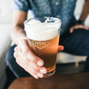 A person holds a Pint Glasses - Set of 4: The Diamond etched with a diamond-shaped Sayers logo.