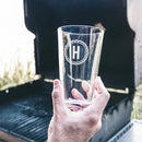 A hand holds a clear pint glass from The Heirloom set by Swanky Badger, featuring a custom circular engraving of the letter H.