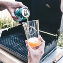 A hand pours beer from a can into a Pint Glasses - Set of 4: The Modern etched with an H emblem, held over a grill.