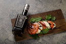 A bottle of Taycte Acetaia Malpighi Platinum IGP – High-Density Balsamic Vinegar of Modena, aged over 25 years, rests beside a Caprese salad of sliced tomatoes, mozzarella, and basil drizzled with the rich balsamic, all arranged on a wooden board atop a gray stone surface.