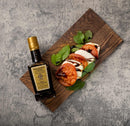 A bottle of Taycte Acetaia Malpighi Gold Balsamic Vinegar of Modena — IGP Certified, Aged 15-20 Years sits beside a Caprese salad on a wooden board atop a gray textured surface.