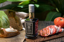 A bottle of Taycte Acetaia Malpighi Platinum IGP – High-Density Balsamic Vinegar of Modena, aged over 25 years, sits on a wooden table next to sliced bread and tomatoes topped with a balsamic drizzle, surrounded by green leaves in natural light.
