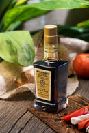 A bottle of Taycte Acetaia Malpighi Gold Balsamic Vinegar of Modena — IGP Certified, Aged 15-20 Years, rests on a wooden table with green leaves, a tomato, prosciutto slices, and vinegar drops nearby.
