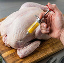 A hand uses the SpitJack SQUIRT 50 Meat Injector Kit with Case and Two Needles, filled with yellow marinade, to inject seasoning into a raw whole chicken resting on a wooden cutting board.