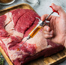 A hand uses the SpitJack SQUIRT 50 Meat Injector Kit with Case and Two Needles to inject marinade into a large piece of raw beef brisket on a wooden cutting board.