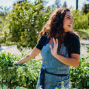 A woman in a blue apron smiles and waves in a lush garden, ready to teach the Messermeister HQ Cooking Class by Messermeister, surrounded by vibrant plants and sunlight.