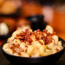 A black bowl of creamy macaroni and cheese, topped with crispy bacon bits and SALTOPIA Cheese Salt: CHEDDAR CHEESE by SALTOPIA Infused Sea Salts, sits on a table with a blurred background.