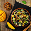A wooden bowl with quinoa salad, black beans, mango, avocado, and toasted seeds sits on a dark cloth. Beside it: sliced mango, a bowl of SALTOPIA Infused Sea Salts' Saltopia Everything Bagel Salt, and a wooden spoon on a rustic table.