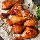 Seven glazed chicken drumsticks on parchment paper, garnished with fresh herbs and parsley, feature a shiny golden-brown coating enhanced by SALTOPIA Bacon Maple Seasoning: JURASSIC PORK infused sea salt.