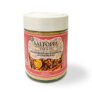 A jar of SALTOPIA Infused Sea Salts Maple Mustard Seasoning Low Sodium: ANOTHER ONE BITES features a red label with roasted vegetables and lemon slices. This low sodium spice comes in a jar with a white lid, ideal for healthy, tasty meals.