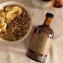 A bowl of oatmeal with bananas and nuts sits beside Under The Bark Syrup Co Bourbon + Vanilla Maple Syrup and a ceramic mug on a beige cloth—an ideal gourmet gift.