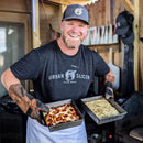 A smiling man in a black Urban Slicer Pizza Worx shirt, gloves, and apron holds two square pans of pizza, proudly showing off his skills with the Urban Slicer Slicer's Special homemade pizza kit in a rustic kitchen.