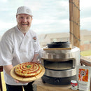 A smiling chef in white holds a pizza with sauce and jalapeños by an outdoor oven. On the table are Urban Slicer Pizza Worx’s Deepest Dish Pi Pack, including homemade deep dish pizza, sauce packet, utensils, and fresh deep dish dough.
