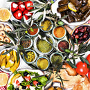 Top-down view of a Mediterranean spread: a central tray featuring the Spice Wheel Mediterranean Spice Wheel by Spice Wheel, filled with vibrant, colorful spices, surrounded by bowls of stuffed peppers, dolmas, olives, yellow peppers, pita bread, tomatoes, salad, and lemon slices—all artfully decorated with fresh olive tree branches.
