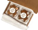 An open cardboard box contains two plastic containers of Texas Pecan Cakes Medium Texas Pecan Cake 2-Pack by Texas Pecan Cakes, each with a round label and a sticker, nestled in protective white packing material—delivering an authentic Texas flavor reminiscent of classic pecan cakes.