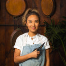 A woman in a white shirt and denim apron stands indoors, smiling as she holds Messermeister Esther Choi Push Button Take-Apart Precision Shears - 7". Copper pans hang on the wooden wall behind her, with a large green plant nearby.