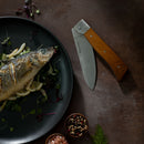 A cooked whole fish with herbs and lemon sits on a dark plate beside the Messermeister Adventure Chef Maple Basecamp Set – 3 Piece, crafted from German stainless steel—ideal for any outdoor cooking set—alongside bowls of peppercorns and pink salt.