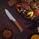 The Messermeister Adventure Chef Maple Basecamp Set - 3 Piece, featuring German stainless steel and a maple handle, rests on a dark surface beside cooked crab with lemon slices, herbs, kitchen shears, a sauce bowl, and scattered spices.