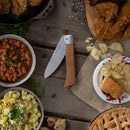 The Messermeister Adventure Chef Maple Basecamp Set - 3 Piece lies open on a rustic table, its wooden handle visible amid plates of baked beans, fried chicken, chips, a hamburger, potato salad, and pie.