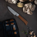 The Messermeister Adventure Chef Maple Basecamp Set – 3 Piece features a German stainless steel chef’s knife with a wooden handle, shown on a dark surface beside oyster mushrooms, herbs, salt, and the outdoor set holding two more knives.