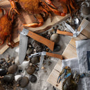 Crabs on a wooden board and sand, surrounded by seashells, rocks, and the Messermeister Adventure Chef Maple Summit Set - 6 Piece; utensils and newspaper complete this inviting outdoor kitchen scene.
