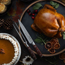 A roasted whole turkey sits on a round slate platter with herbs, cranberries, and dried oranges beside the Messermeister Avanta Pakkawood Knife Block Set - 10 Piece, carving knives, pumpkin pie with whipped cream, pinecones, and fall leaves on a dark table.