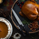 A roasted turkey on a round tray with herbs and dried orange slices, carved with two knives from the Messermeister Avanta Damascus Block Set - 10 Piece, sits beside pumpkin pie, cranberries, and autumn leaves on a dark table.
