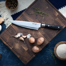 A Messermeister Avanta Damascus Starter Set - 3 Piece chef’s knife rests on a wooden cutting board with garlic, small potatoes, salt, and thyme. Nearby on a blue tablecloth are bowls of peppercorns, flour, and a few eggs.