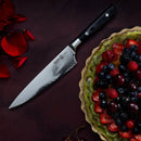 A Messermeister Avanta Damascus Starter Set - 3 Piece chef’s knife rests on a dark surface beside a scalloped plate of mixed berries, with red rose petals nearby, highlighting the elegance of Messermeister craftsmanship.