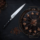A glossy chocolate cake with chocolate decorations sits on a dark plate beside a sharp Messermeister Avanta Damascus knife from the 10-piece block set. Chocolate shavings are scattered around the cake and knife on the tabletop.