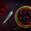 A fruit tart topped with mixed berries sits on a dark surface beside the Messermeister Avanta Damascus Block Set knife, scattered rose petals, cherries, and a strawberry.