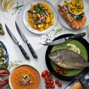 A table set with seafood—whole fish on green sauce, seafood soup, oysters, prawns, gnocchi, herbs, cherry tomatoes, crusty bread—and the Messermeister Avanta Damascus Fine Edge Steak Knife Set (4 Piece) for elegant dining.