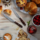 A breakfast scene with croissants, a muffin, raspberries, a latte, a strawberry dessert drink, and two Messermeister Avanta Pakkawood knives from the 10-piece set on a light wooden table. Pastries and crumbs are scattered around.