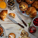 A breakfast spread with croissants, muffins, berries, granola, orange juice, coffee, and two Messermeister Avanta Pakkawood Steak Knives on a light wooden table. One croissant is sliced open by the fine-edge Messermeister blade; crumbs scatter nearby.