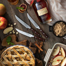Overhead view of a wooden table with an apple pie, the Messermeister Avanta Pakkawood Knife Block Set (10 Piece) by Messermeister, a bottle of Maker’s Mark bourbon, brown sugar in a black cup, sliced apples, cinnamon sticks, apple chunks, and a white dish with apple slices.