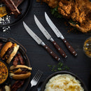 The Messermeister Avanta Pakkawood Knife Block Set (10 Piece) features stainless steel knives with stylish pakkawood handles, beautifully arranged amidst plates of fried chicken, burgers, ribs, mashed potatoes, a whiskey glass, fork, and herbs.