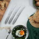 The Messermeister Avanta Stainless Fine Edge Steak Knife Set (4-piece) is arranged on a white countertop beside pita bread, a green cloth, a bowl of greens with a sunny-side-up egg, a fork, and a cutting board with pita in the upper left.