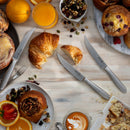A breakfast scene with pastries, croissant, orange slices, mixed nuts, coffee with latte art, orange juice, and a set of Messermeister Avanta Stainless Fine Edge Steak Knives (4 Piece) for steakhouse-quality precision on a light wooden table.