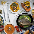 A table set with seafood—whole fish in green sauce, shrimp with mango salsa, oysters, lobster, and more—is ready to enjoy using the Messermeister Avanta Stainless Fine Edge Steak Knife Set (4 Piece) for precise cuts and effortless dining.