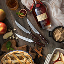 Overhead view of the Messermeister Avanta Pakkawood Steak Knife Set (4-piece) with fine-edge blades and pakkawood handles on a wooden table, surrounded by Maker’s Mark bourbon, apples, apple pie, brown sugar, cinnamon sticks, a drink, and sliced fruit.