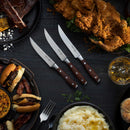 The Messermeister Avanta Pakkawood Steak Knife Set (4 Piece) sits at the center of a table laden with fried chicken, burgers, mashed potatoes, barbecue ribs, and a glass of whiskey.