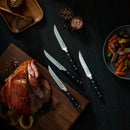 A Messermeister Avanta Steak Knife Set – 4 Piece, with ergonomic handles, is arranged on a dark textured surface alongside a wooden board of sliced roast meat, mixed vegetables, a honey jar with dipper, and bowls of salt and pepper.